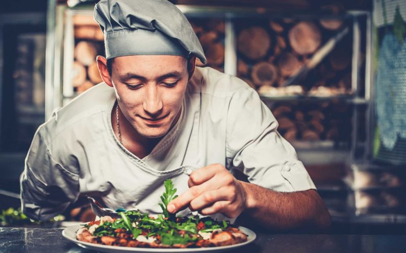 Food concept. Preparing traditional italian pizza. Young smiling chef in white uniform and gray hat decorate ready dish with green rucola herbs in interior of modern restaurant kitchen. Ready to eat.